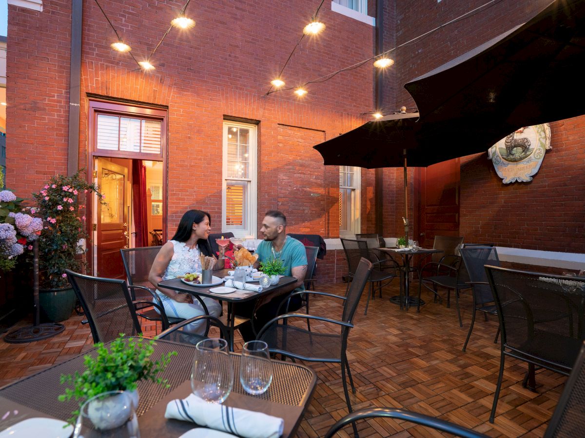 A couple is dining outdoors at a restaurant patio with string lights, surrounded by tables, chairs, and potted plants, on a brick plaza.