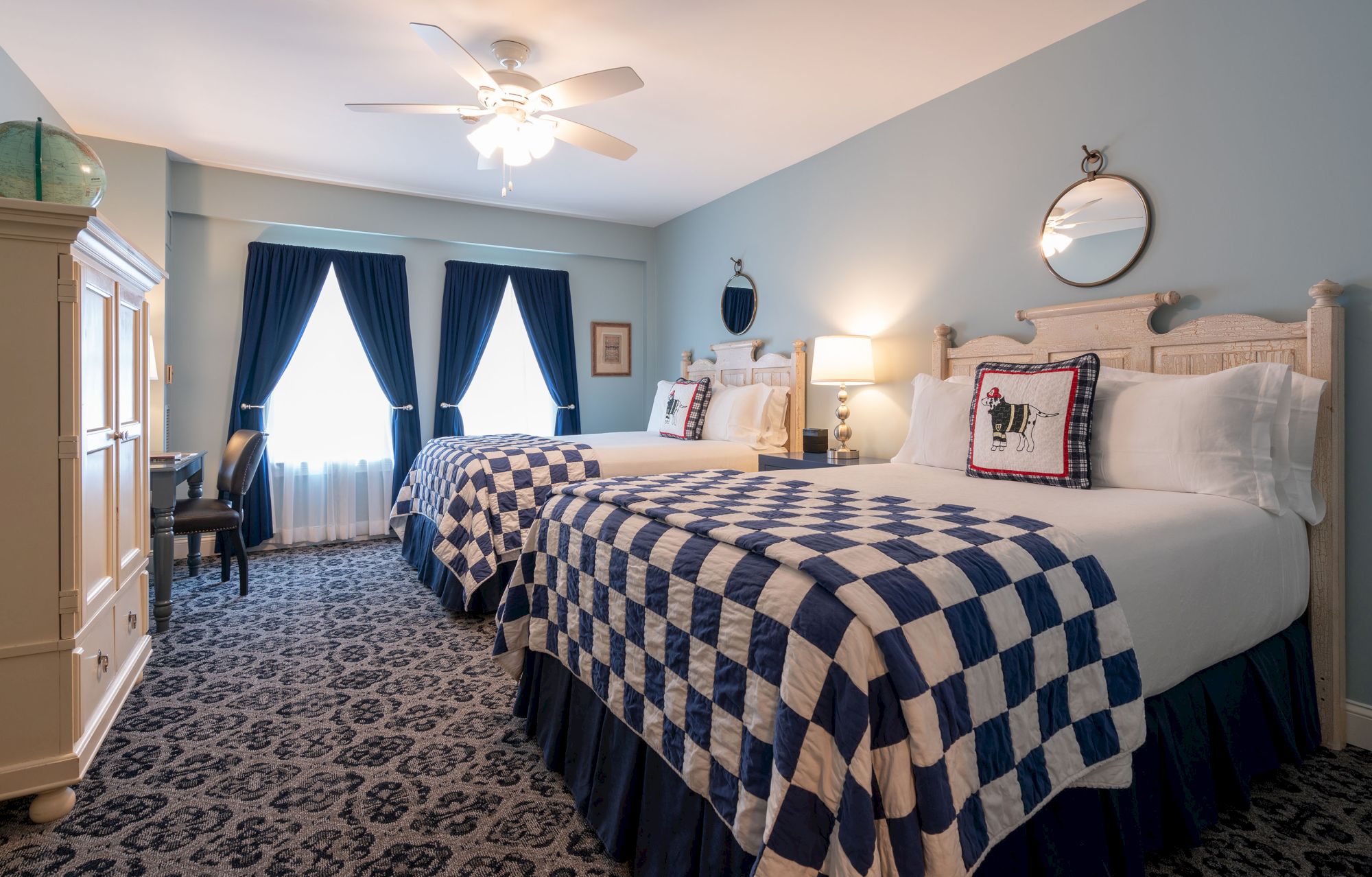 A hotel room with two beds, blue checkered bedspreads, blue curtains, a ceiling fan, and a lamp on a bedside table.