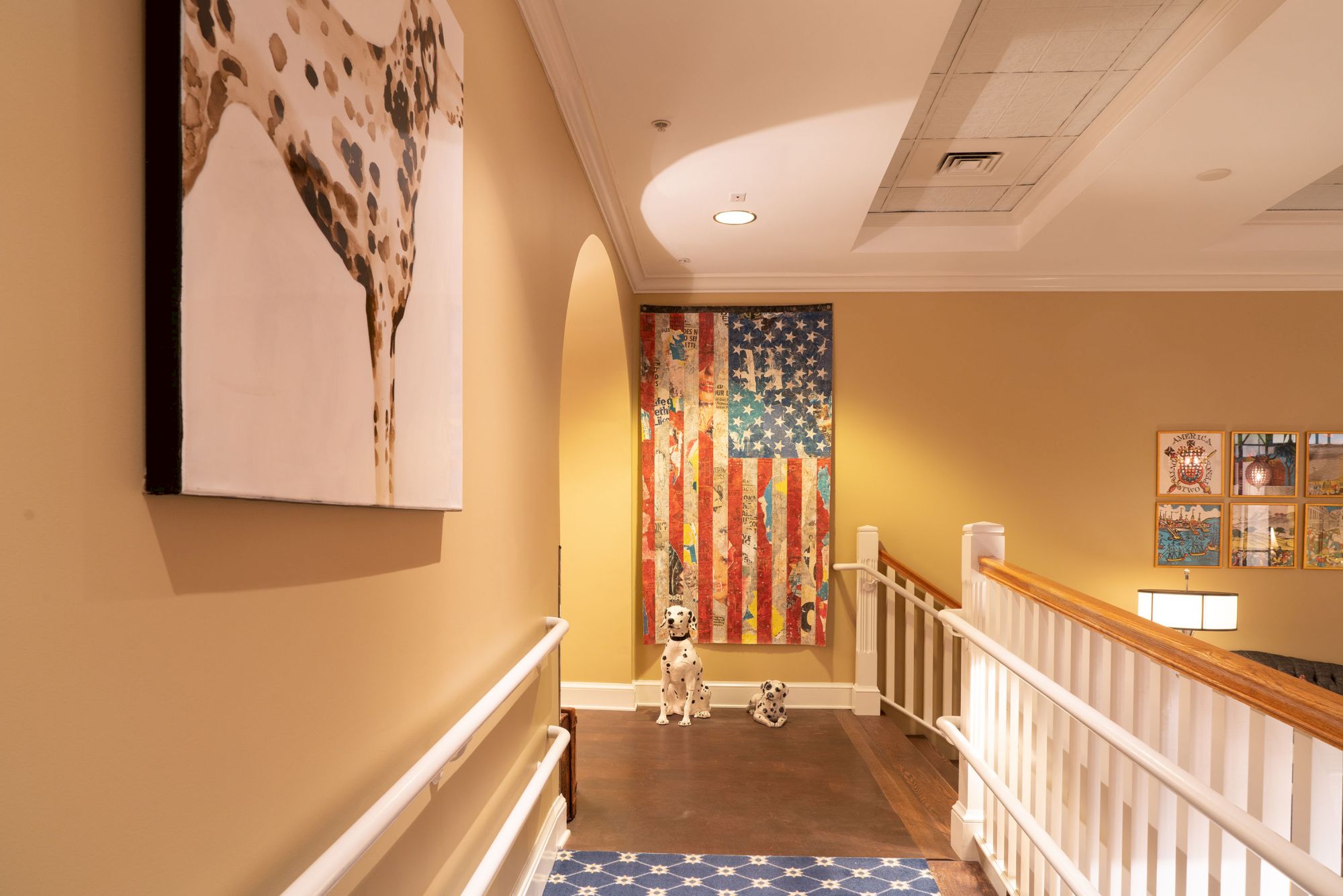 A hallway with dog-themed decor, including a painting, statue, and American flag art. The walls are beige, and there is a railing along the side.