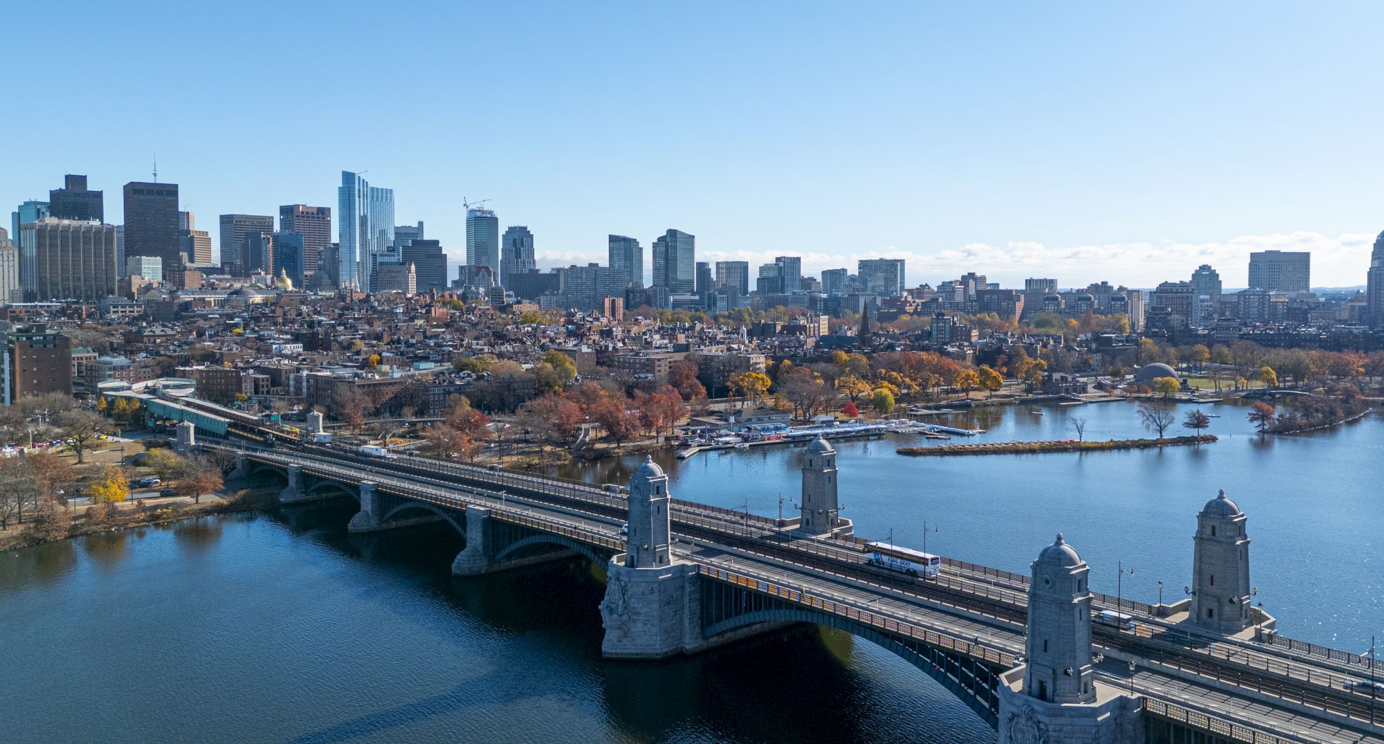 A city skyline with a bridge over a river in the foreground, surrounded by autumn trees and urban buildings.