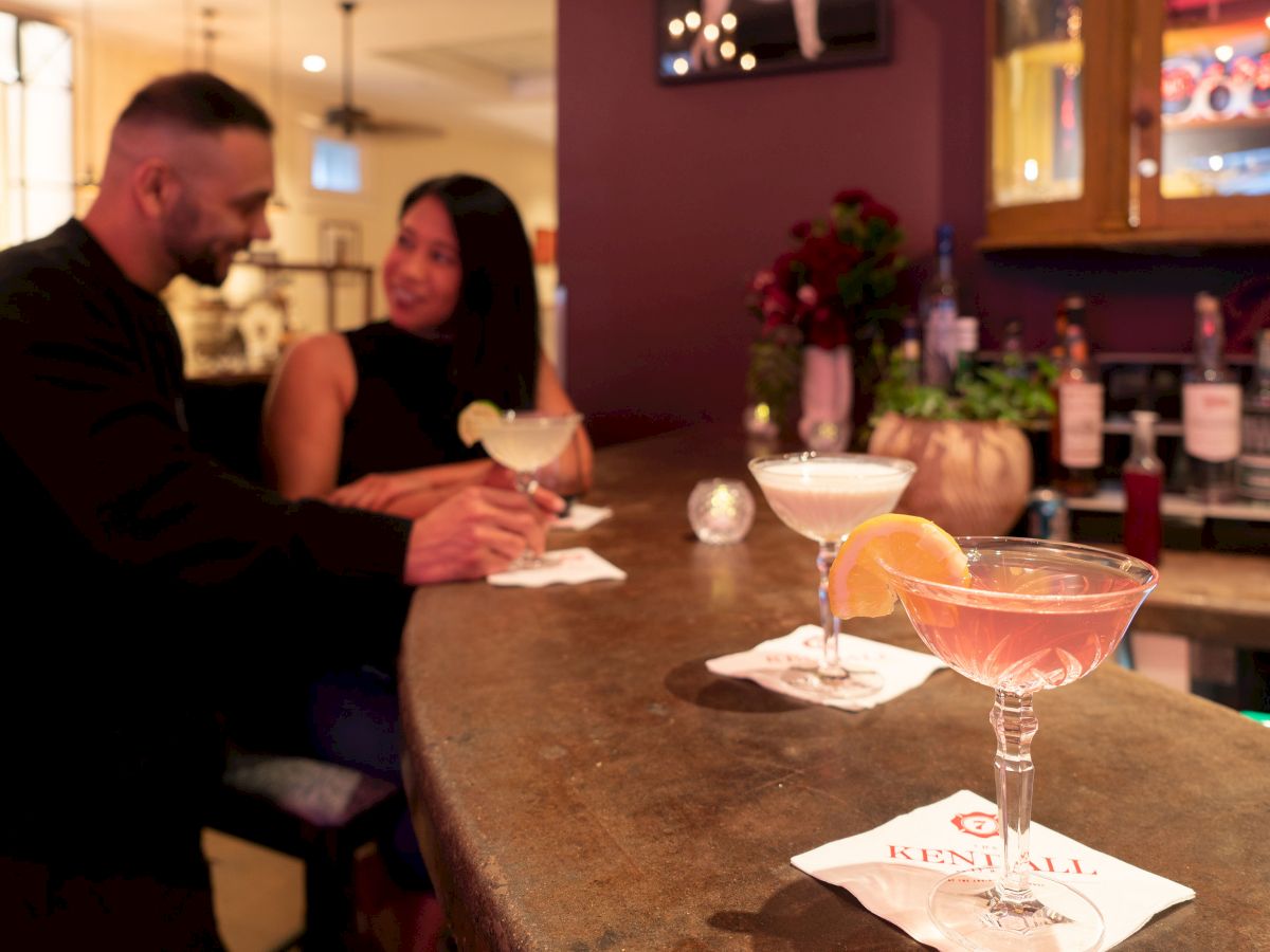 A man and a woman are sitting at a bar, chatting while holding cocktails. Three other cocktails are placed on the bar counter, garnished with fruit slices.