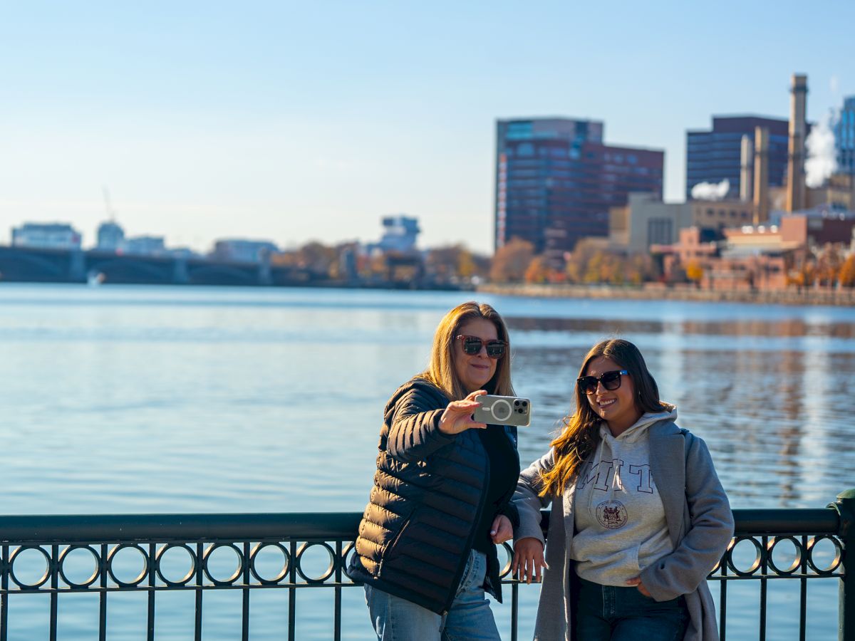 Two people taking a selfie by a waterfront with city buildings in the background, both wearing sunglasses.