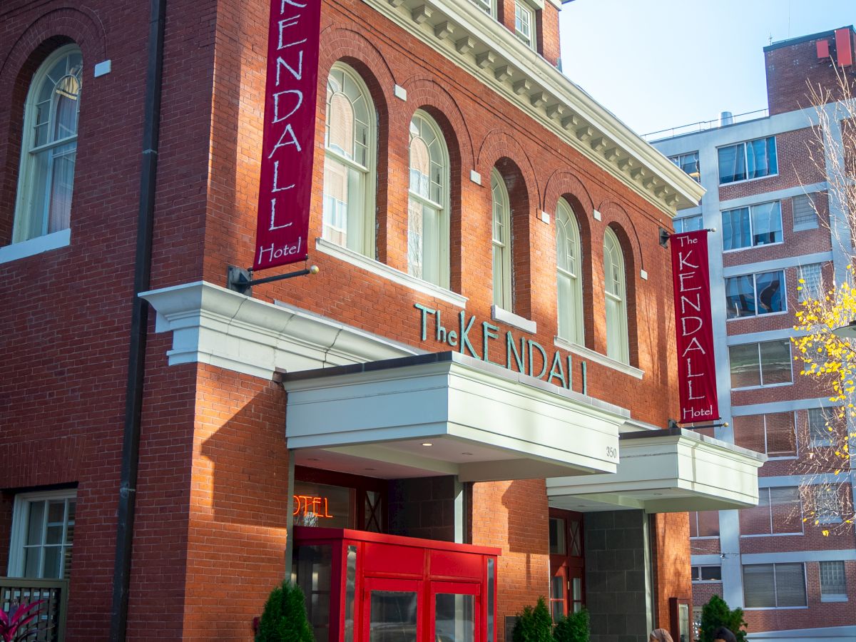 The image shows a red-brick building labeled "The Kendall Hotel" with a red entrance, plants, and bicycles nearby.