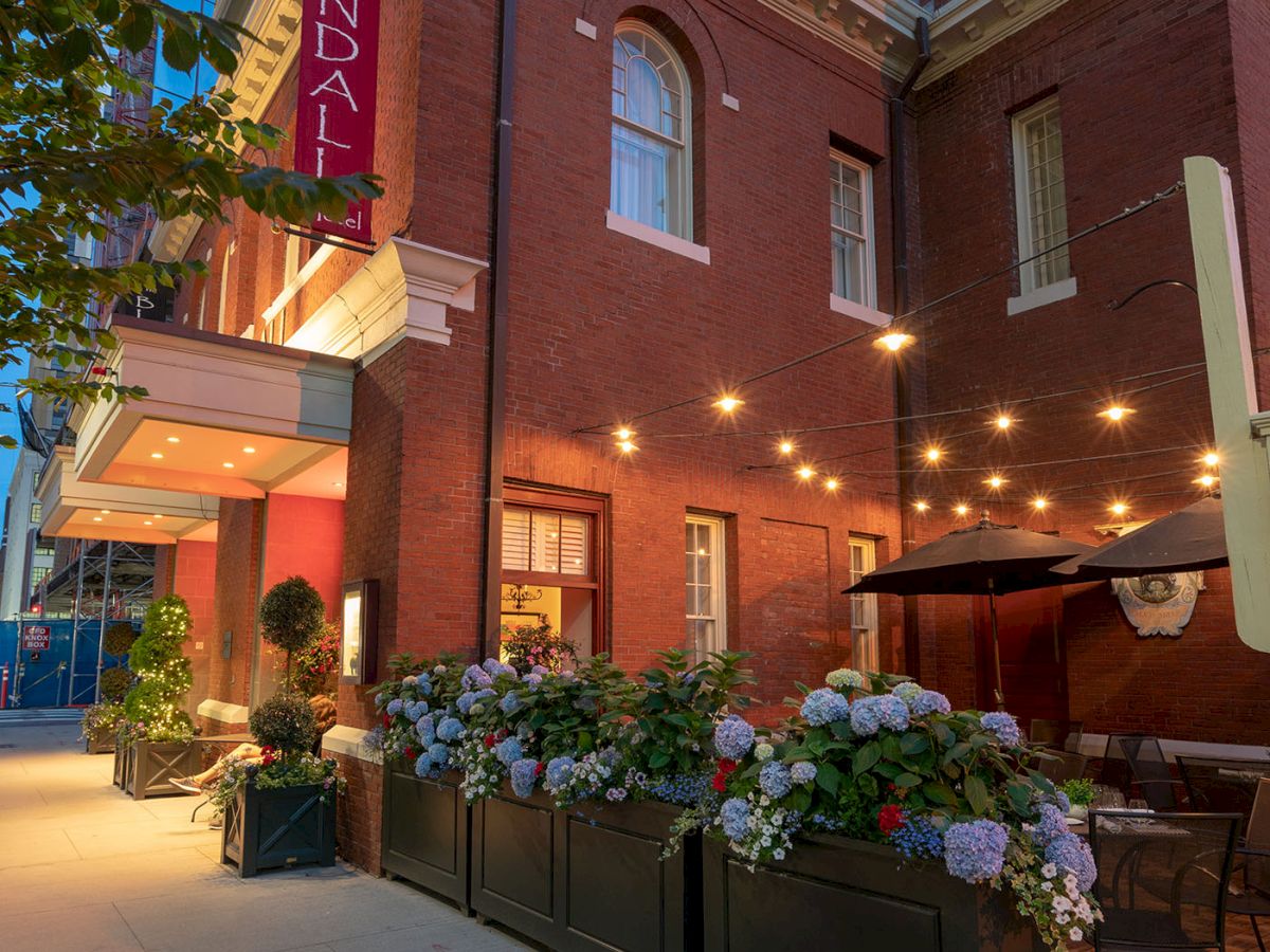 A charming outdoor restaurant scene with string lights, flowers, brick building, trees, and a red sign that reads 