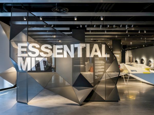 The image shows a modern exhibit with the words "ESSENTIAL MIT" displayed on a geometric structure in an art gallery or museum setting.