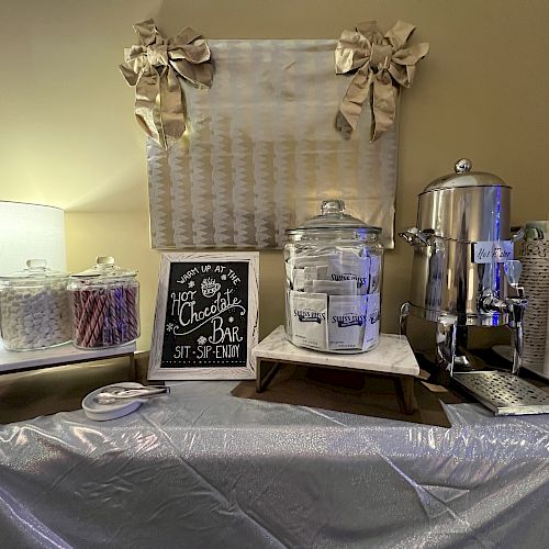 A hot chocolate bar setup with a sign, jars of ingredients, a dispenser, mugs, and napkins on a table covered with a silver cloth.