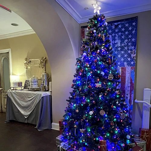 A decorated Christmas tree with blue lights and presents underneath, set in a room with festive decorations and a table in the background.