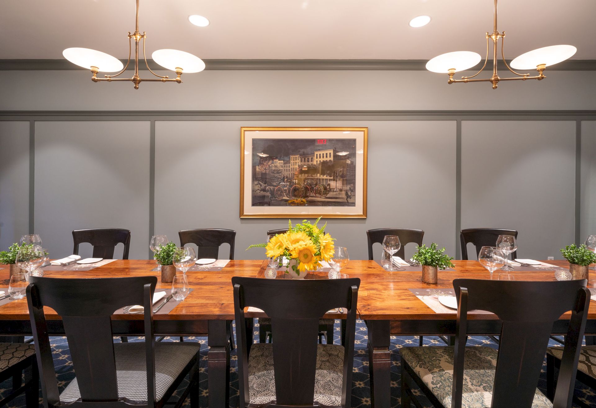 A dining room with a wooden table, black chairs, a flower centerpiece, wall artwork, and hanging lights. There are plants and table settings.
