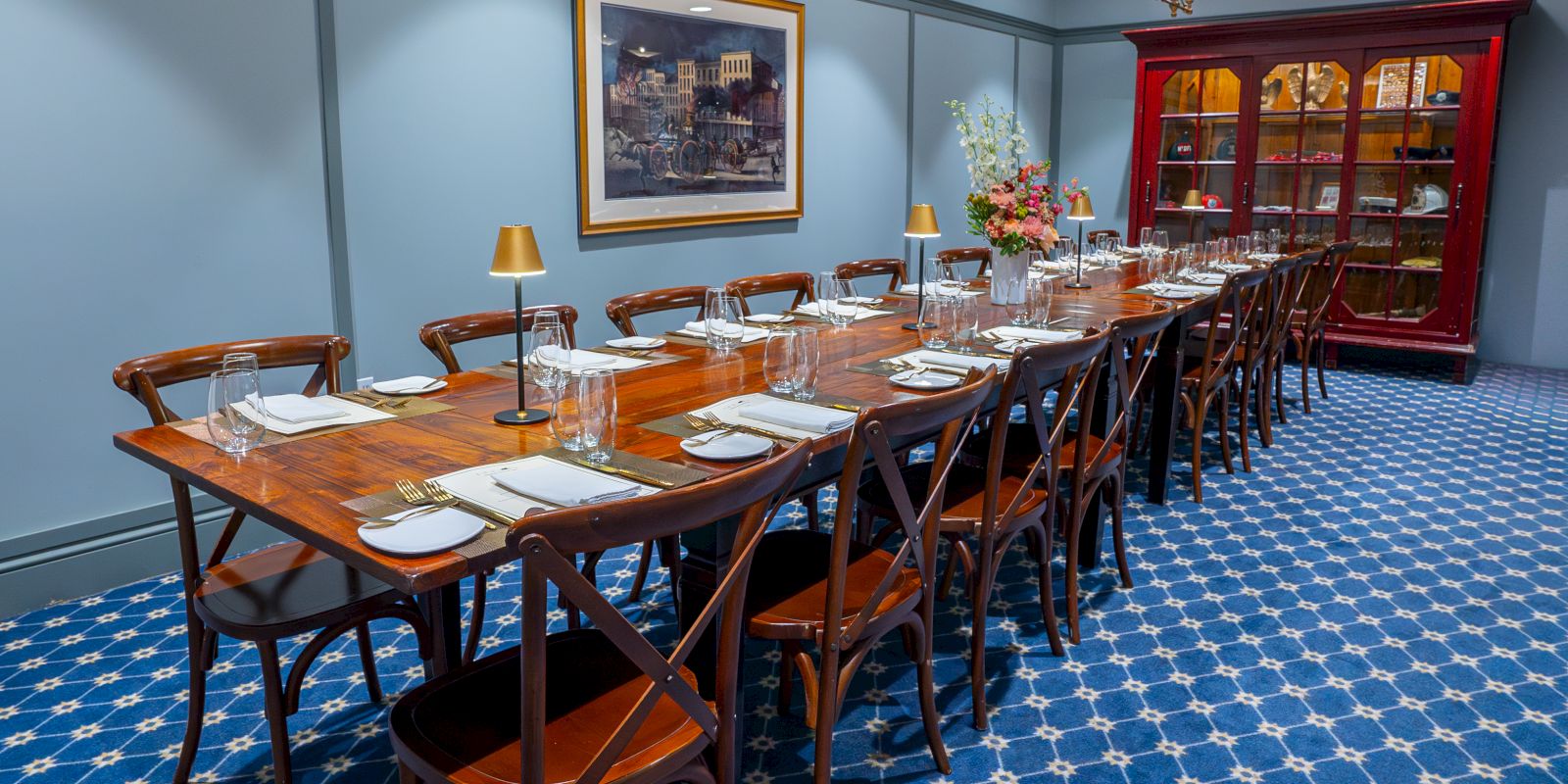 A long wooden dining table set for a formal meal with plates, glasses, cutlery, and napkins arranged for many guests in a blue-walled room.