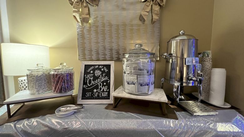 A hot chocolate bar setup with jars, a drink dispenser, a metallic urn, and a decorative sign and bows.