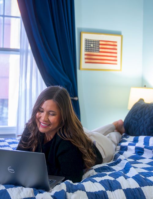 A person is lying on a bed in a blue-themed room, using a laptop. There is a framed flag on the wall and curtains by the window.