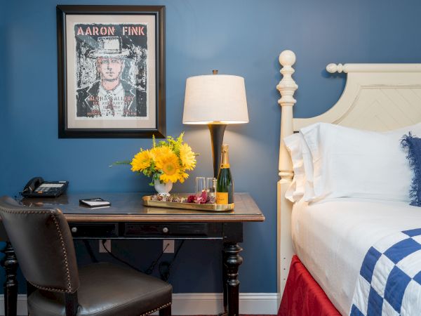 A cozy room with blue walls and white furniture, featuring a framed picture, a desk with a lamp, flowers, champagne, and a bed with a checkered blanket.