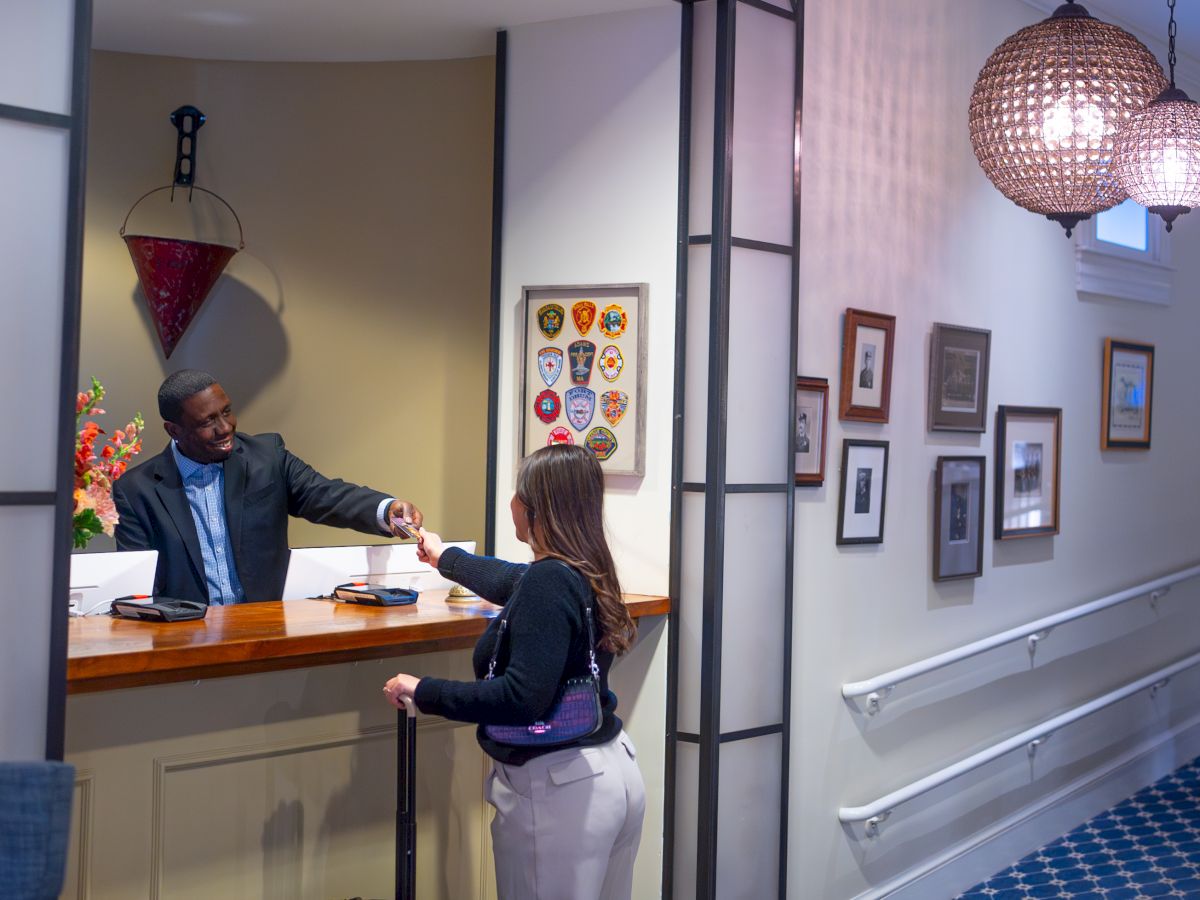 A person is checking into a hotel at the reception desk. The hallway is decorated with framed art and blue carpeting.