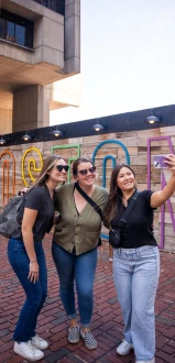 Three friends take a selfie in front of a colorful sign that spells &ldquo;B O S T O N&rdquo; on a brick plaza, smiling and posing together.
