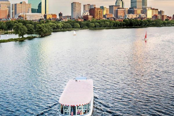 A boat is cruising on a river with a city skyline and sailboat in the background under a partly cloudy sky.