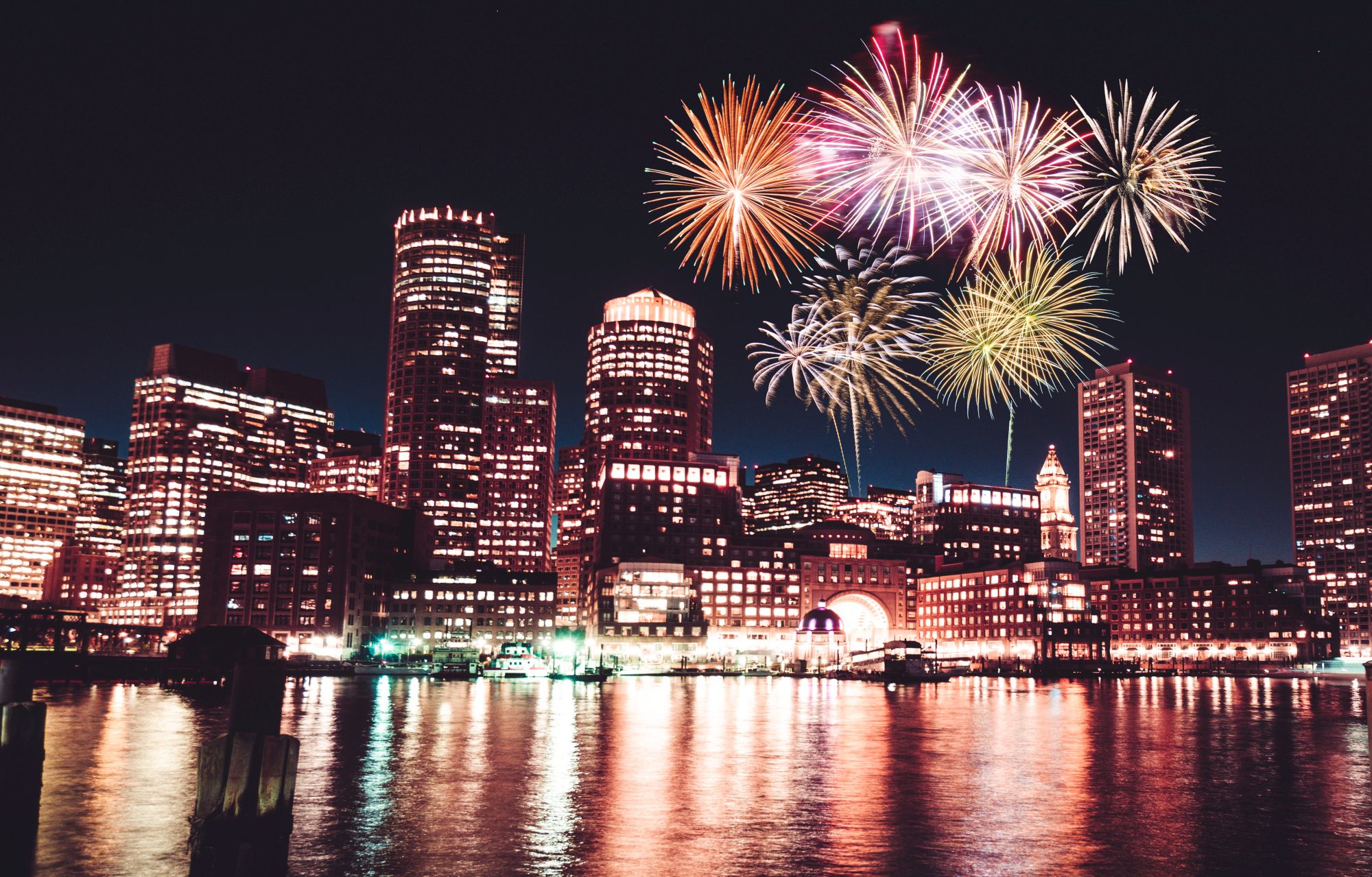 A city skyline at night with colorful fireworks lighting up the sky, reflecting in the water below.
