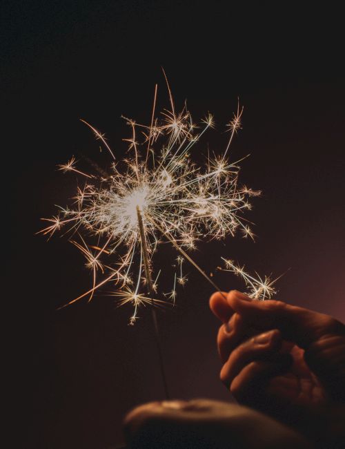 A hand holds a sparkling lit sparkler against a dark background, with small bursts of light illuminating the scene.