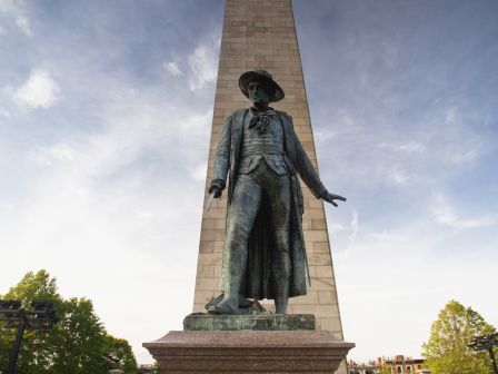 A statue of a historical figure in front of an obelisk, set against a partly cloudy sky with trees in the background.