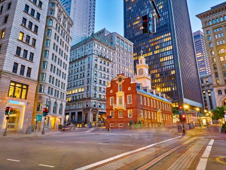 An historic brick building is surrounded by modern skyscrapers in a cityscape, blending old and new architecture as night falls.