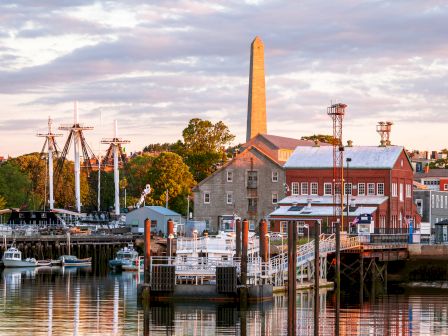 The image shows a scenic harbor with historical ships, dockside buildings, and an obelisk monument under a cloudy sky.