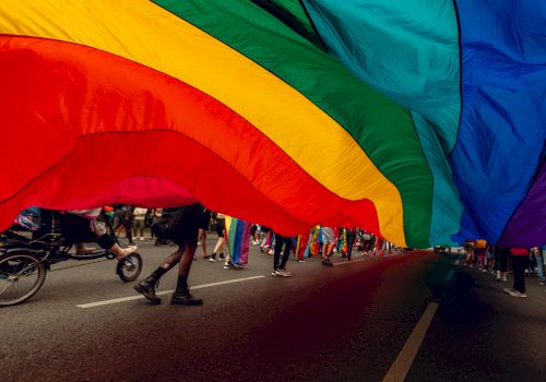 Large rainbow flag held above people walking on a street during a parade or event, capturing the vibrant colors and movement.