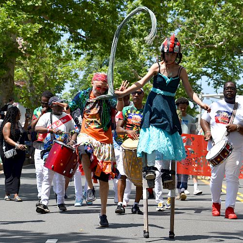 A lively street parade with performers on stilts and musicians, including drummers, marching down a tree-lined road.