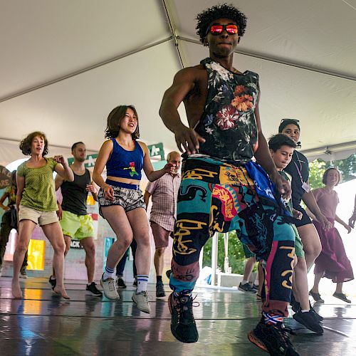 A group of people dancing energetically under a tent, led by a person in colorful attire and sunglasses.