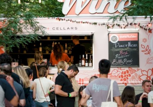 People are gathered at a wine truck with a chalkboard menu, surrounded by greenery, enjoying a social outdoor event.