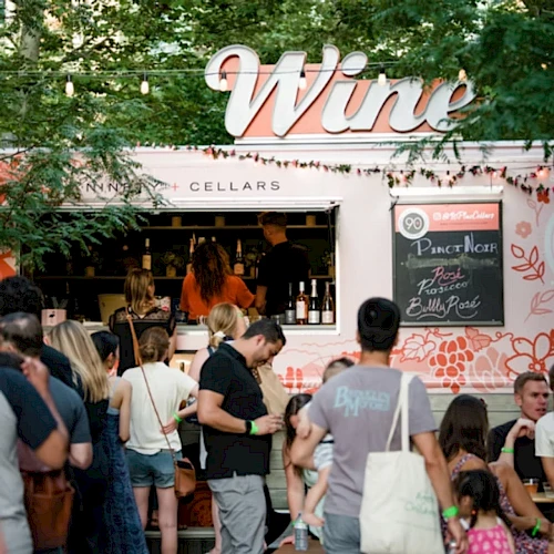 People are gathered at a wine truck with a chalkboard menu, surrounded by greenery, enjoying a social outdoor event.