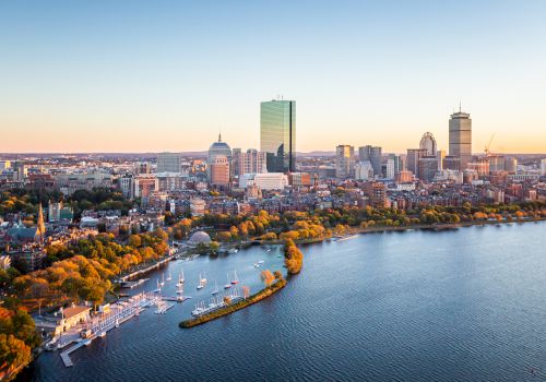 Aerial view of a cityscape with a prominent river and skyline during sunset, featuring boats and lush greenery along the riverbank.