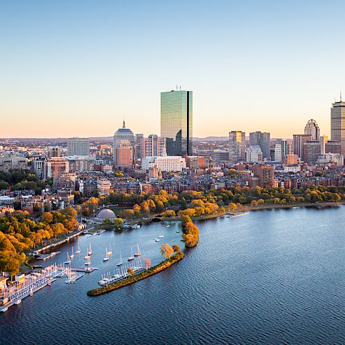 Aerial view of a cityscape with a prominent river and skyline during sunset, featuring boats and lush greenery along the riverbank.