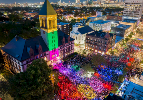 A vibrant night scene shows a large crowd gathering outside an illuminated building with colorful lights in a city setting.