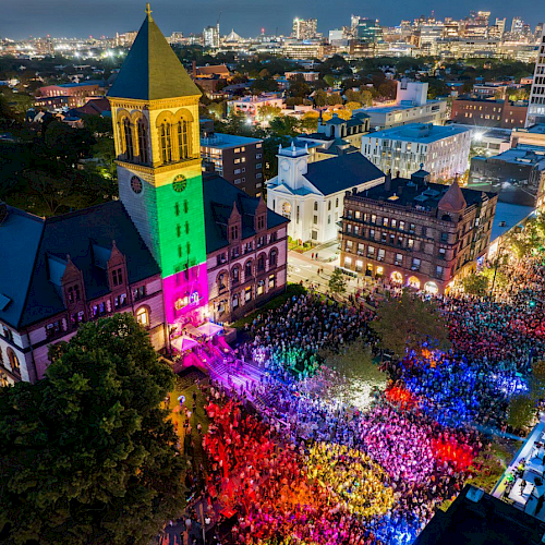 A vibrant night scene shows a large crowd gathering outside an illuminated building with colorful lights in a city setting.