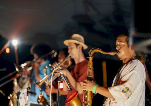Musicians playing brass instruments on stage, with focus on saxophonist in front; others blurred. Dimly lit performance setting.