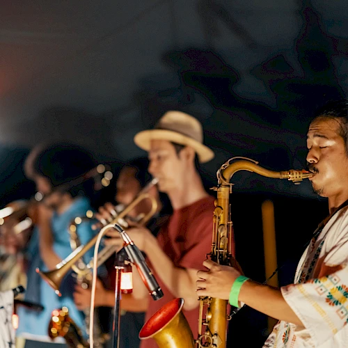 Musicians playing brass instruments on stage, with focus on saxophonist in front; others blurred. Dimly lit performance setting.