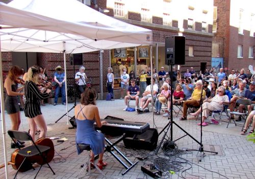 A band performs under a white tent in a street, with a large seated audience watching. Musical instruments include a keyboard, cello, and violin.