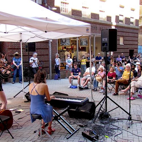 A band performs under a white tent in a street, with a large seated audience watching. Musical instruments include a keyboard, cello, and violin.