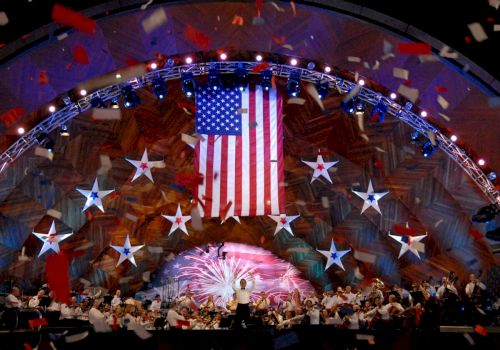 A concert stage with an orchestra, a large American flag, stars, and falling confetti, suggests a festive, patriotic celebration.