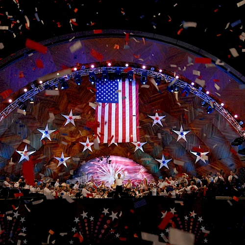 A concert stage with an orchestra, a large American flag, stars, and falling confetti, suggests a festive, patriotic celebration.