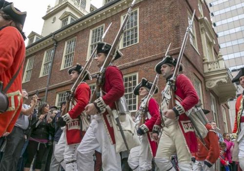 The image shows a group of people in historical military uniforms, marching in front of an old brick building, likely part of a parade or reenactment.