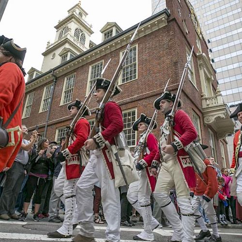 The image shows a group of people in historical military uniforms, marching in front of an old brick building, likely part of a parade or reenactment.