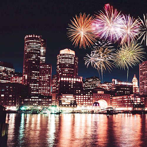 A city skyline at night with fireworks bursting in the sky, reflected on the water.