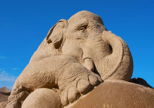 A detailed sand sculpture of an elephant under a clear blue sky is depicted in this image.