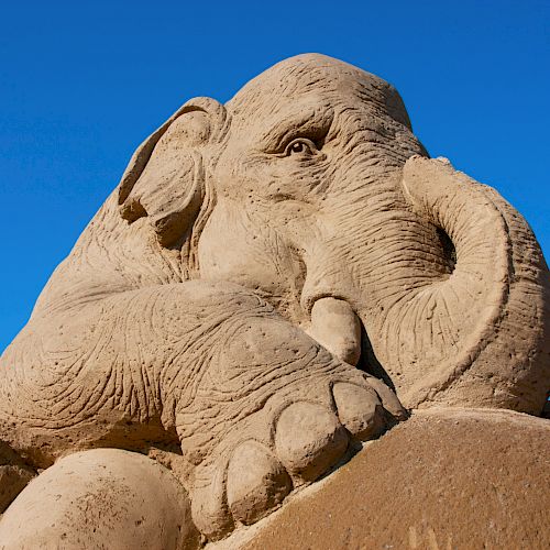 A detailed sand sculpture of an elephant under a clear blue sky is depicted in this image.