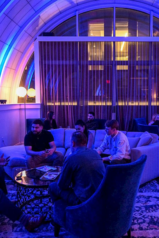 A lounge with people sitting on sofas under blue lighting in a spacious, curved room with large windows at the back.