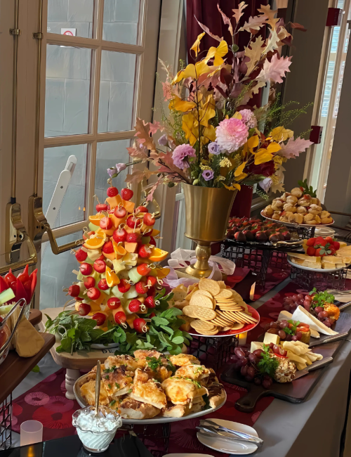 A buffet table with a variety of appetizers, fresh vegetables, sandwiches, and a decorative flower centerpiece by a window.