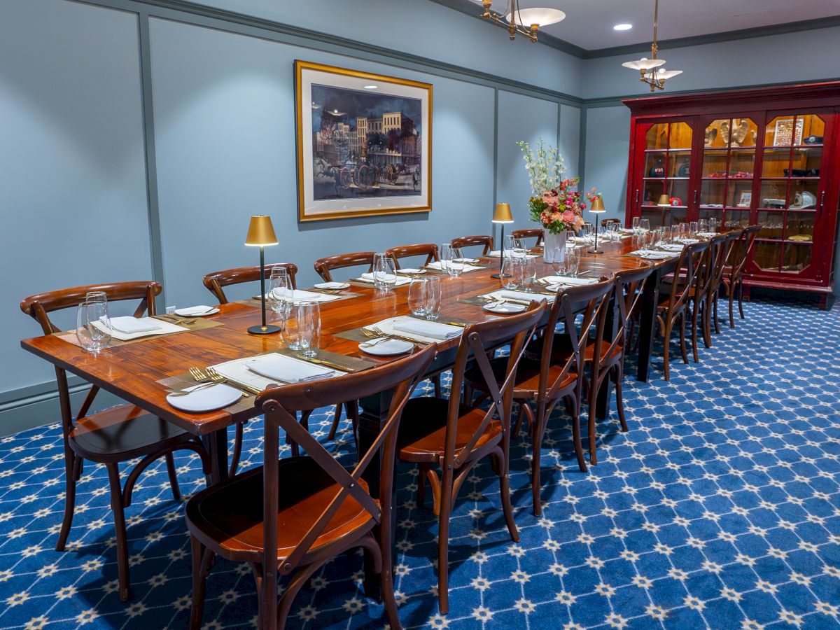 A long dining table set for a meal with elegant tableware, against blue walls, featuring a painting, china cabinet, and floral arrangement.