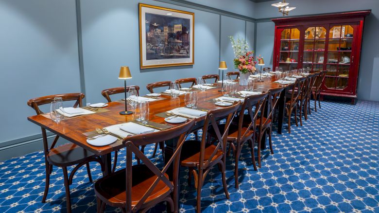 A long dining table set for a meal with elegant tableware, against blue walls, featuring a painting, china cabinet, and floral arrangement.
