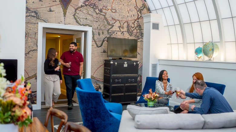 A group of people is socializing in a stylish room with a world map wall, blue chairs, and a cozy atmosphere.