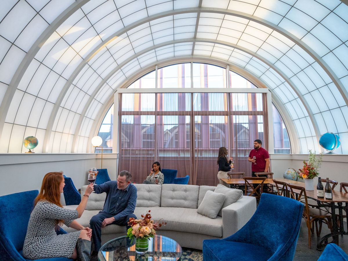 The image shows a bright room with a curved ceiling, people conversing on couches, and standing at a table, featuring globes and modern decor.